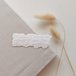 May include: A sticker with the text "Oh Jesus I surrender myself to you. Take care of everything" in a handwritten style. The sticker is placed on a beige book, with dried flowers in the background.