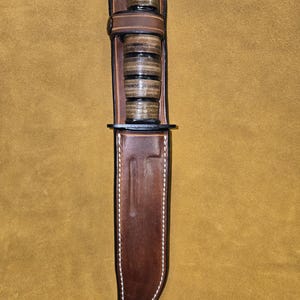 May include: A brown leather knife sheath with a matching handle and a black top. The handle has a stacked appearance with alternating dark and light brown layers. The sheath is stitched with white thread.