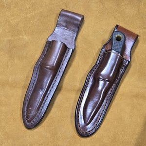 Peut inclure: Deux étuis en cuir marron, dont un avec un couteau partiellement visible. Les étuis sont cousus sur les bords et ont une boucle en haut. Le fond est de couleur beige.