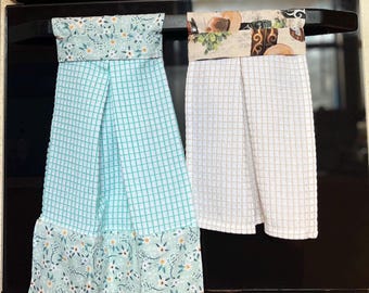 May include: Two hanging kitchen towels. One is light blue with a grid pattern and floral trim. The other is white with a grid pattern and a patterned top with images of food. Both are hanging on a black oven handle.