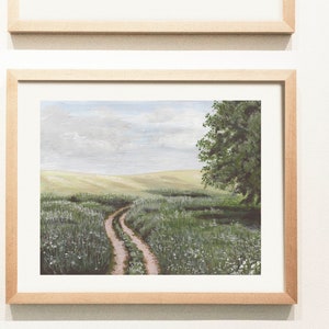 May include: A framed painting of a dirt road leading through a field of green grass and white flowers. A single tree stands on the right side of the road, with a blue sky and white clouds in the background.