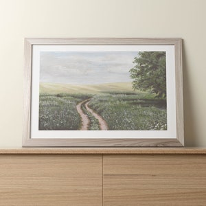 May include: A framed painting of a winding dirt road through a field of green grass and white wildflowers. A single tree stands on the right side of the road.