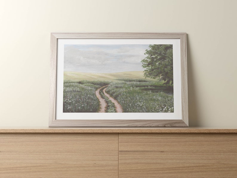 May include: A framed painting of a winding dirt road through a field of green grass and white wildflowers. A single tree stands on the right side of the road.