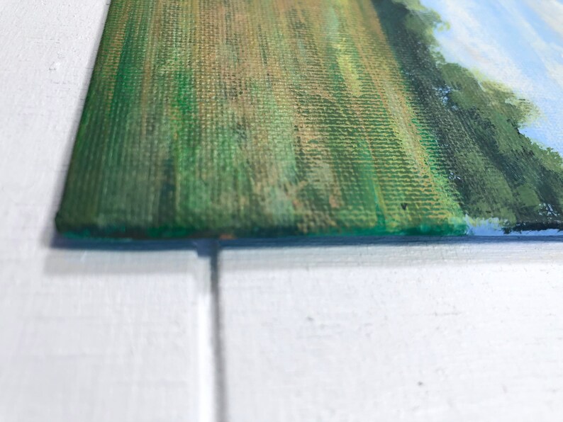May include: Close-up of a green and brown canvas painting with a textured surface. The painting depicts a landscape with a field of grass and trees in the background.