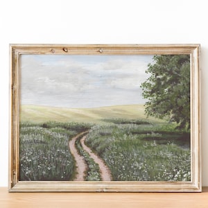 May include: A framed painting of a dirt road winding through a field of green grass and white wildflowers. The road leads to a distant hill and a lone tree stands on the right side of the painting. The sky is a light blue with white clouds.