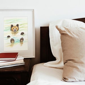 May include: A framed watercolor painting of a brown bear swimming in a blue and green lake. The bear's head is above the water and its paws are visible below the surface. The painting is on a white background and is framed in a white frame.