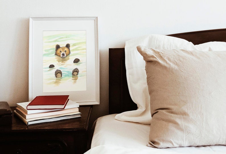 May include: A framed watercolor painting of a brown bear swimming in a blue and green lake. The bear's head is above the water and its paws are visible below the surface. The painting is on a white background and is framed in a white frame.