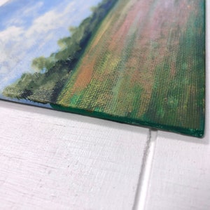 May include: A small painting on canvas depicting a landscape scene with a blue sky, white clouds, and a green field with a hint of orange and pink.