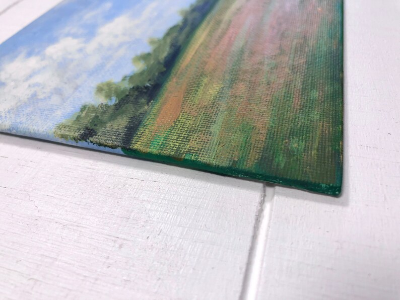 May include: A small painting on canvas depicting a landscape scene with a blue sky, white clouds, and a green field with a hint of orange and pink.
