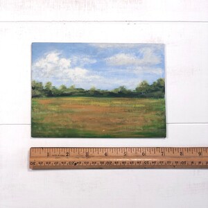 May include: A small painting depicting a landscape with a field of green grass, a brown dirt road, and a blue sky with white clouds.