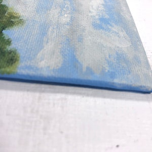 May include: Close-up of the bottom edge of a small canvas painting. The painting depicts a blue sky with white clouds and a green tree on the left side.