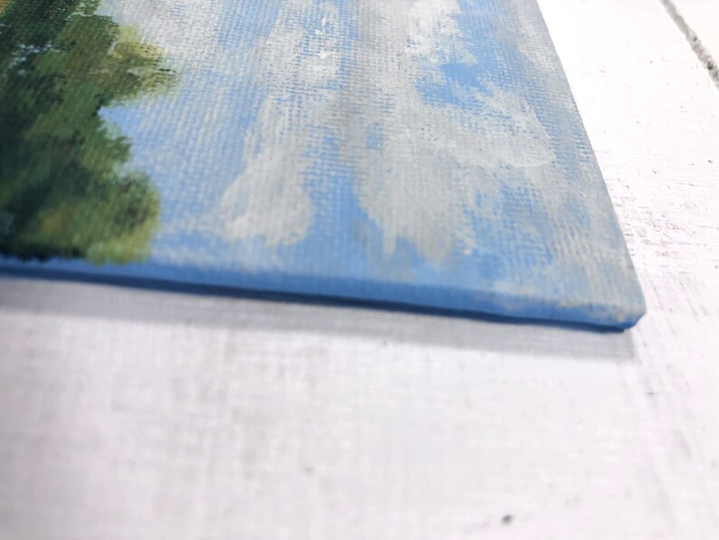 May include: Close-up of the bottom edge of a small canvas painting. The painting depicts a blue sky with white clouds and a green tree on the left side.