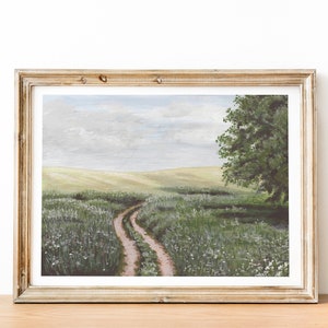 May include: A framed painting of a dirt road winding through a field of green grass and white wildflowers. The road leads to a large green tree in the distance. The sky is a light blue with white clouds.