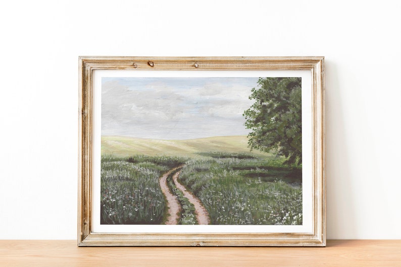 May include: A framed painting of a dirt road winding through a field of green grass and white wildflowers. The road leads to a large green tree in the distance. The sky is a light blue with white clouds.