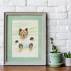 May include: A watercolor painting of a brown bear floating in a body of water. The bear's head is above the water, and its paws are visible below the surface. The painting is framed in a light gray frame with a green mat.