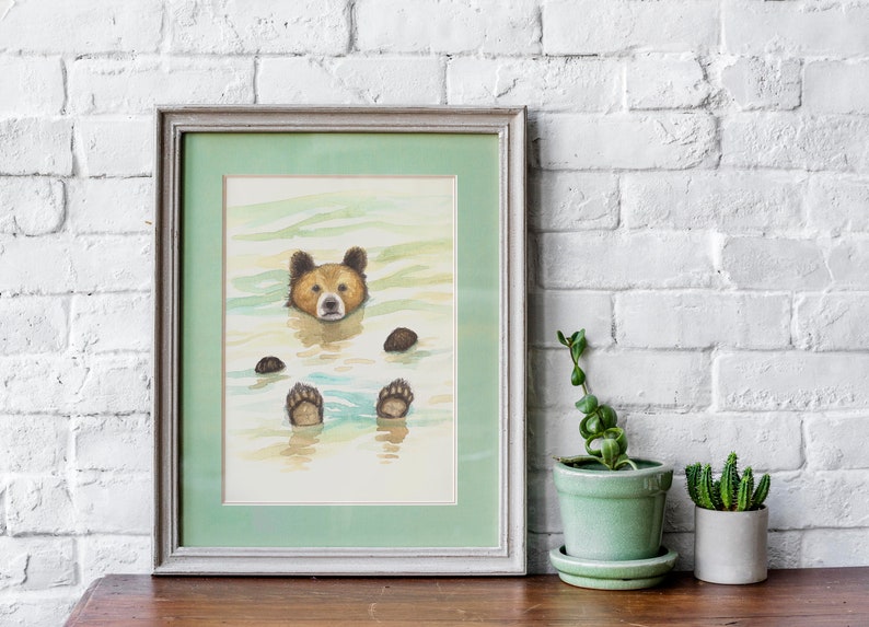 May include: A watercolor painting of a brown bear floating in a body of water. The bear's head is above the water, and its paws are visible below the surface. The painting is framed in a light gray frame with a green mat.