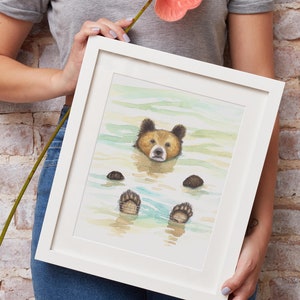 May include: A framed watercolor painting of a brown bear swimming in a light blue body of water. The bear's paws are visible above the water. The painting is in a white frame.