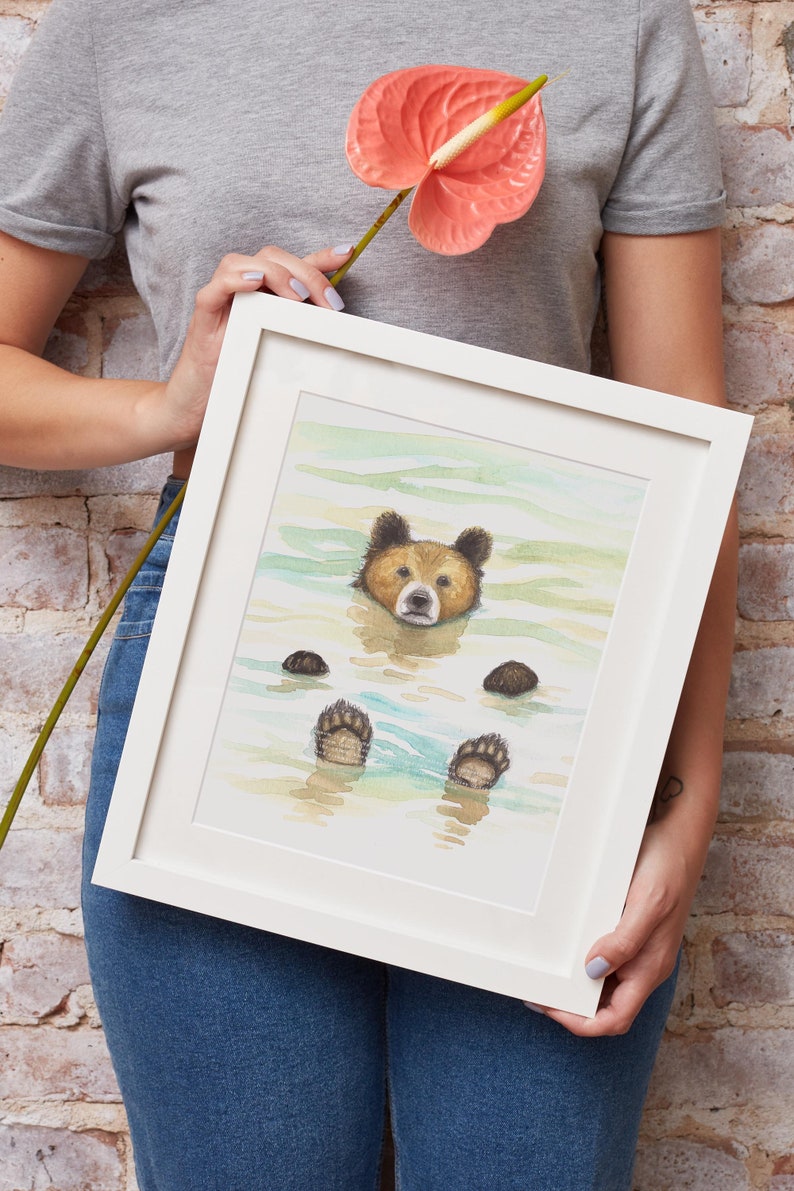 May include: A framed watercolor painting of a brown bear swimming in a light blue body of water. The bear's paws are visible above the water. The painting is in a white frame.