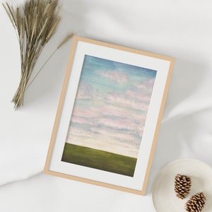May include: A framed painting of a landscape with a green field and a pink and blue sky. The frame is made of light wood.