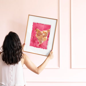 Puede incluir: Una mujer sostiene una impresi&oacute;n enmarcada de una gallina marr&oacute;n sobre un fondo rosa. El marco es dorado y la impresi&oacute;n est&aacute; en papel texturizado.