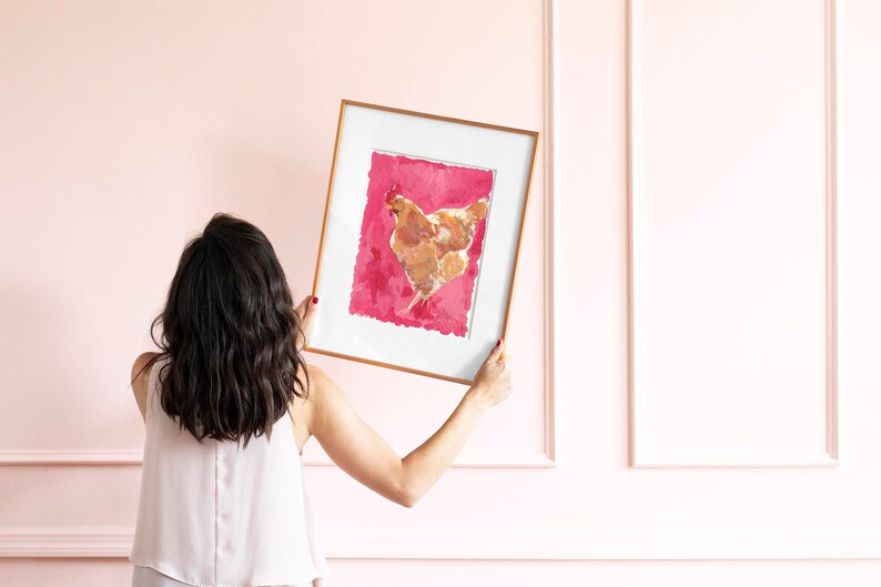 Puede incluir: Una mujer sostiene una impresi&oacute;n enmarcada de una gallina marr&oacute;n sobre un fondo rosa. El marco es dorado y la impresi&oacute;n est&aacute; en papel texturizado.