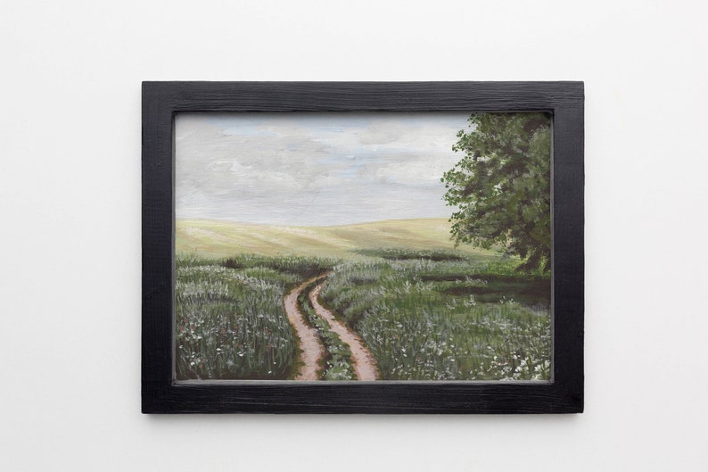 May include: A framed painting of a dirt road winding through a field of green grass and white wildflowers. The sky is cloudy and a single tree stands on the right side of the road.