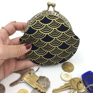 May include: A small, round coin purse with a gold and navy blue fabric exterior. The purse has a decorative metal clasp and a scalloped edge. The fabric features a repeating pattern of gold scales on a navy blue background.
