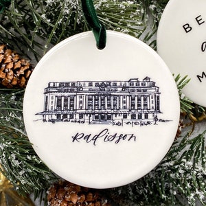 May include: White ceramic Christmas ornament with a black and white line drawing of a building with the text "Radisson" underneath.