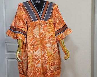 May include: An orange, long-sleeved dress with a V-neck and a leaf-like pattern. The dress has a ruffled neckline and sleeve cuffs, with blue and brown striped accents. The dress is displayed on a mannequin with a gold head and arms.