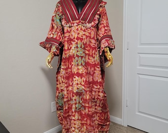 May include: A full-length, red patterned dress with a V-neckline and long sleeves. The dress features a floral-like pattern in shades of yellow, green, and blue. The neckline and sleeves are trimmed with a contrasting burgundy fabric and gold accents.
