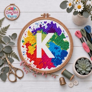 Könnte beinhalten: Ein runder Holzstickrahmen mit einem Kreuzstichmuster, das einen großen weißen Buchstaben K zeigt, zentriert vor einem lebendigen, mehrfarbigen Hintergrund im Aquarellstil aus roten, gelben, grünen, blauen und violetten Stichen.