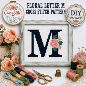 May include: A cross stitch pattern featuring a large dark navy letter M decorated with pink and peach flowers and green leaves, framed in a light blue wooden frame. Includes embroidery thread spools, scissors, and a thimble on a white surface.