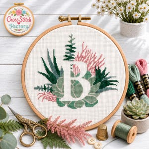 May include: A circular wooden embroidery hoop containing a cross-stitch design of the letter B surrounded by various green succulents and pink floral elements on white fabric. The design features shades of dark green, sage, and soft pink.