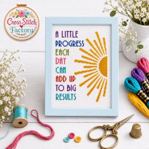 Peut inclure: Une broderie au point de croix encadrée représentant un soleil jaune et le texte A LITTLE PROGRESS EACH DAY CAN ADD UP TO BIG RESULTS en lettres multicolores. L'ouvrage est présenté dans un cadre bleu clair sur un support en bois blanc.