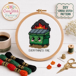 Puede incluir: Un proyecto de punto de cruz en un bastidor de madera que muestra un contenedor verde con fuego encima y el texto EVERYTHING'S FINE. El diseño está bordado sobre tela blanca. La imagen incluye hilo de bordar, tijeras y botones con forma de llama.