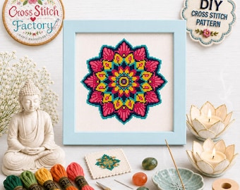 May include: A framed cross stitch mandala pattern featuring vibrant pink, yellow, and teal geometric shapes on white fabric. The display includes embroidery floss, a small pair of gold scissors, a needle, a Buddha statue, and lotus candle holders.