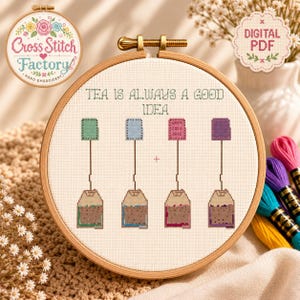 May include: A circular wooden embroidery hoop containing a cross-stitch pattern on white fabric. The design features four tea bags in green, blue, pink, and purple, with the text "TEA IS ALWAYS A GOOD IDEA" and "WARM COZY DAYS" stitched above.