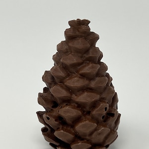 May include: A brown, 3D printed pine cone ornament. The ornament is detailed with a textured surface and has a small hole at the top for hanging.