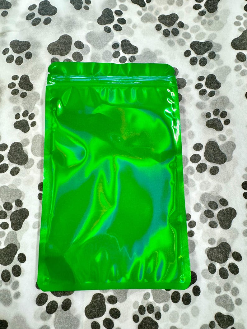 May include: A green iridescent resealable plastic bag with a zipper closure. The bag is lying flat on a white surface with a black paw print pattern.