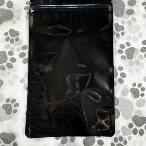 May include: A black, stand-up pouch with a zipper closure. The pouch is made of a shiny, metallic material.