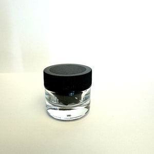 May include: A small, clear glass jar with a black lid. The jar is filled with a dark, crumbly substance.