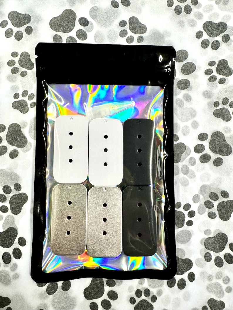 May include: A package of six blank metal dog tags in three different colors: white, silver, and black. The tags are rectangular with rounded corners and have four holes for attaching a chain or other hardware.