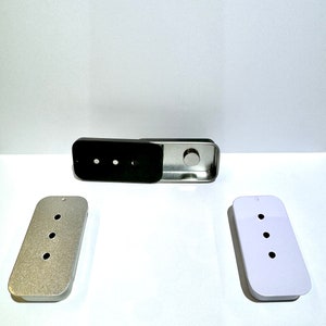 May include: Three metal cases with three holes each. The cases are silver, white, and black. The black case is open, revealing a silver interior.