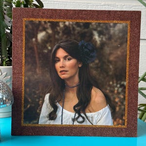 May include: A framed portrait of a woman with long dark hair, wearing a white off-the-shoulder top and a blue flower in her hair. The frame has a brown glittery border with a gold inner edge. The background is a blurred outdoor scene.