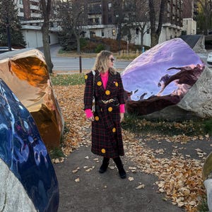 Può includere: Una donna indossa un lungo cappotto a quadri con bottoni a forma di faccina gialla e un maglione rosa. Il cappotto è nero, rosso e verde. Indossa stivali neri e occhiali da sole. Lo sfondo include grandi sculture riflettenti colorate e foglie autunnali.