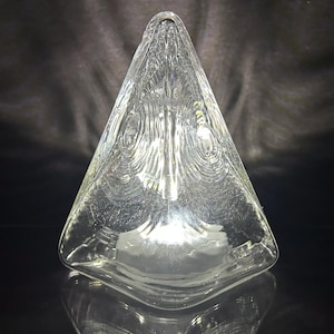 May include: Clear glass triangular decorative object. The object has a textured surface with a pattern of concentric circles. The base is a flat, square shape. The object is displayed against a dark background.