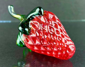 May include: A handcrafted glass strawberry sculpture. The sculpture features a vibrant red body with a textured surface, mimicking the fruit's natural appearance. The stem and leaves are a glossy green, adding contrast. The artwork is a decorative piece.