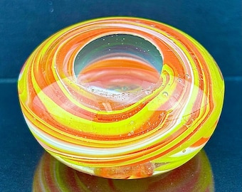 Könnte beinhalten: Ein runder Briefbeschwerer aus Glas mit einem wirbelnden Muster aus leuchtendem Orange, hellem Gelb und Weiß. Der klare Glaskörper hat eine glatte, polierte Oberfläche mit einer kreisförmigen Vertiefung an der Oberseite und kleinen Luftblasen.