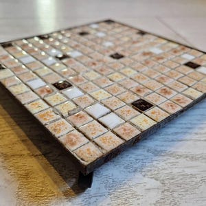 May include: Square trivet with a mosaic tile design. The tiles are a mix of beige, white, and brown, arranged in a grid pattern. The trivet has a dark metal frame and small, curved feet.