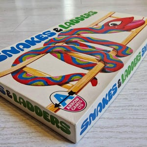 May include: A rectangular board game box titled Snakes & Ladders by Arrow Games Ltd. The white box features a colourful illustration of a red, blue, and green patterned snake winding through a wooden ladder. The title is printed in blue and green.
